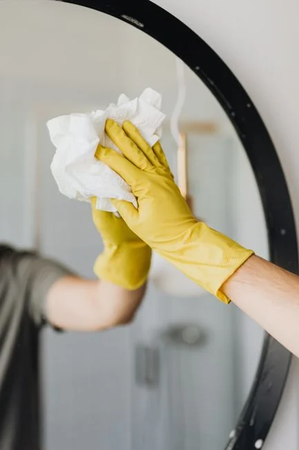 services-04 Side view of a young woman in latex gloves, concentrating as she cleans a mirror in a bright room during the day